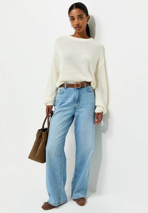 Vrouw staand, gekleed in een witte gebreide trui, lichtblauwe wijde jeans, bruine riem, bruine platte schoenen en een bruine handtas vasthoudend.