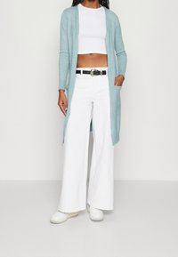 Jasnoniebieski dziergany kardigan z długimi rękawami, otwarty z przodu, z dwoma bocznymi kieszeniami, w połączeniu z białym krótkim topem i szerokimi białymi spodniami.