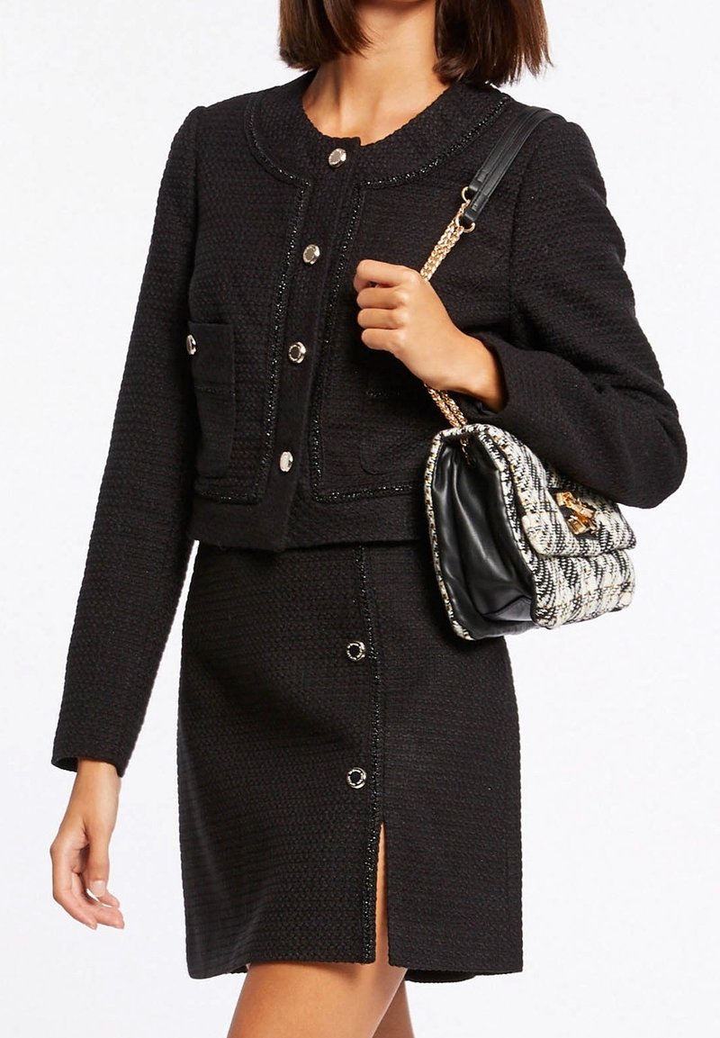 Musta tweed-takki pyöreällä pääntiellä ja napituksella, yhdistettynä vastaavaan hameeseen, jossa on etumellinen halkio ja napit. Mukana teksturoitu käsilaukku.