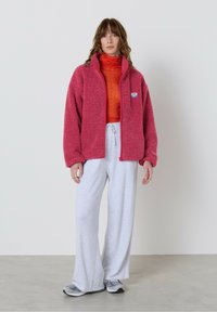 Rožnato rožnat plišast pulover z zadrgo spredaj in majhnim logotipom, združen s svetlo rdečim žabljim ovratnikom ter sivimi širokimi trenirkami in sivimi supergami.