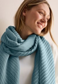 Écharpe bleu clair avec un design texturé en côtes. Tissu doux, enroulé lâchement autour du cou, mettant en valeur sa forme ondulée et ses plis superposés.