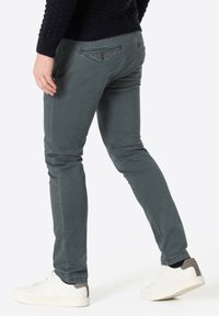 Schlanke, dunkelgrüne Baumwollhosen mit strukturiertem Stoff, Gesäßtaschen und einem kleinen braunen Knopf-Akzent, kombiniert mit weißen Sneakers.