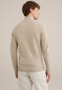 Beige pulóver, strukturált kötésű, álló gallérral és bordás aljjal, világos színű nadrággal párosítva. Az anyag finom fényű.