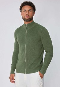 Homme avec des cheveux bouclés courts et une barbe, portant un pull vert zippé et un pantalon blanc, se tenant avec une main dans la poche.