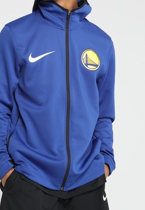 Osoba ubrana w niebieską sportową kurtkę zapinaną na zamek z logo Golden State Warriors i znakiem Nike, ręce w kieszeniach, na gładkim białym tle.
