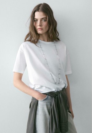 Junge Frau mit langen braunen Haaren trägt ein weißes T-Shirt, helle Jeans aus Denim und eine grau gestreifte Jacke, die um ihre Taille gebunden ist, sowie eine lange silberne Halskette.