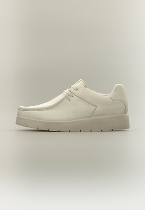 Chaussure décontractée basse blanche avec texture en cuir lisse, bout arrondi, fermeture à lacets et semelle beige épaisse sur un fond neutre.