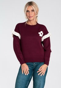Kobieta z blond włosami, nosząca bordowy sweter z białymi falbankami na rękawach i białym wzorem kwiatu, w połączeniu z niebieskimi dżinsami.