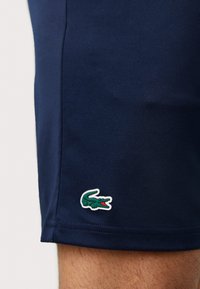 Σκούρες μπλέ σορτς από λεία ύφανση με λευκή περιθωριοποίηση και πράσινο λογότυπο κροκοδείλου στο στρίφωμα, σε ίσια γραμμή και χωρίς επιπλέον διακοσμήσεις.