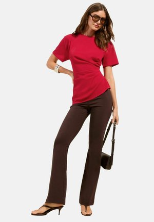 Žena nosiaca červený krátky rukáv, tmavohnedé nohavice, slnečné okuliare a sandále, držiaca čiernu kabelku s jednou rukou na bokoch.