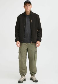Veste en polaire noire avec col haut, t-shirt gris en dessous, pantalon cargo vert avec poches, et chaussures de randonnée beiges.