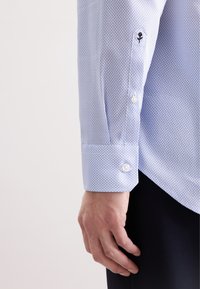 Camicia azzurra chiara con un motivo geometrico, dotata di bottoni bianchi e una rosa ricamata sulla manica. Tessuto dal tocco morbido.