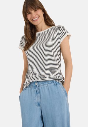 Femme souriante aux cheveux bruns portant un t-shirt à manches courtes noir et blanc à rayures et un pantalon ample bleu clair, les mains dans les poches.