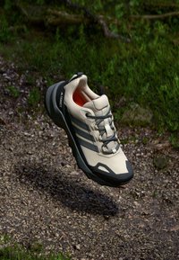 Zapatilla de trail running en color beige claro con acentos grises, parte superior texturizada, cierre de cordones y suela negra. Presenta un interior naranja contrastante.