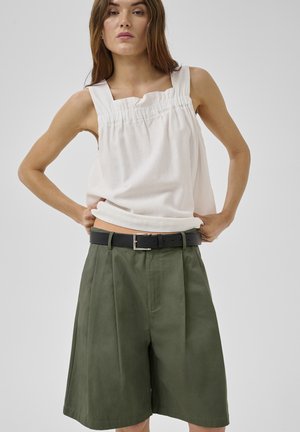 Woman wearing a sleeveless white top and high-waisted olive green wide-leg shorts with a black belt, standing against a plain background.