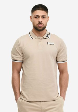 Mann mit kurzen dunklen Haaren und Bart, trägt ein beiges Lonsdale London Polohemd mit gestreiftem Kragen und Ärmeln, Hände in den Taschen, neutraler Hintergrund.