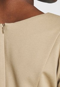 Beżowa bluzka z szerokim dekoltem, wykonana z teksturowanej tkaniny. Posiada dyskretny zamek z tyłu do zapięcia.