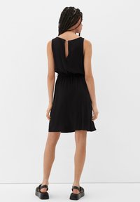 Frau mit langem geflochtenem Haar, die ein ärmelloses schwarzes Kleid mit einem schlüssellochförmigen Ausschnitt auf der Rückseite und schwarze Plateausandalen trägt, und mit dem Rücken zur Kamera steht.