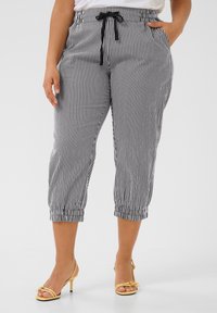 Pantalons cropped à rayures noires et blanches avec des poignets élastiques, une ceinture à cordon de serrage et des poches latérales. Le matériau semble léger et texturé.