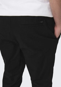 Pantalones negros con una tela texturizada, que cuentan con un bolsillo trasero y una pequeña etiqueta de la marca. El diseño es de corte recto con un estilo minimalista.