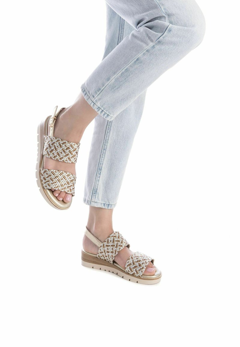 Sandali intrecciati beige e oro con due ampie cinghie, una suola imbottita e un design con cinturino posteriore, su uno sfondo chiaro.