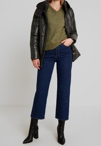 Chaqueta acolchada negra, suéter de pico verde olivo, jeans acampanados oscuros y botines de tacón negros. Texturas suaves y corte a medida.