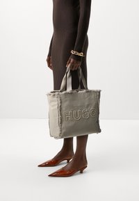 Grau canvas Tragetasche mit fransigen Kanten und besticktem "HUGO"-Schriftzug. Verfügt über doppelte Stoffgriffe. Braunes geripptes Kleid und spitze Schuhe.