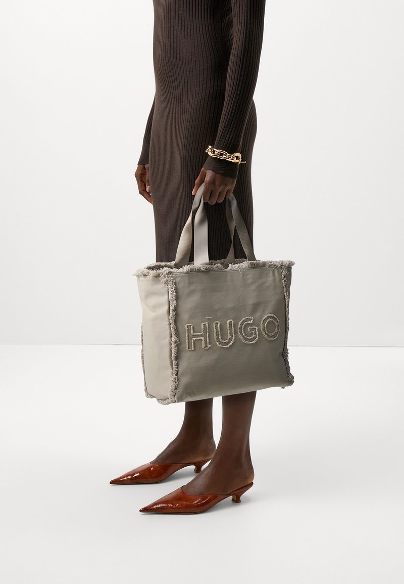 Grau canvas Tragetasche mit fransigen Kanten und besticktem "HUGO"-Schriftzug. Verfügt über doppelte Stoffgriffe. Braunes geripptes Kleid und spitze Schuhe.