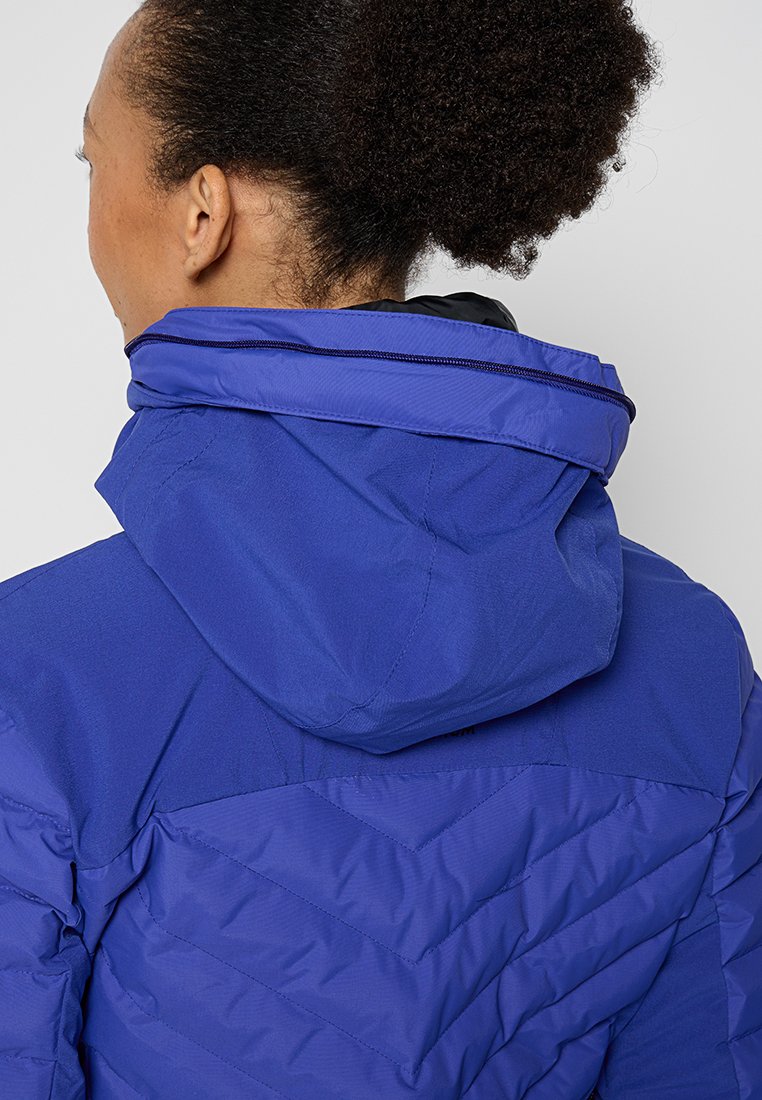 Veste à capuche violette avec un design texturé et matelassé. Elle présente un col montant et une capuche ajustable, mettant en avant des détails de couture contrastants.