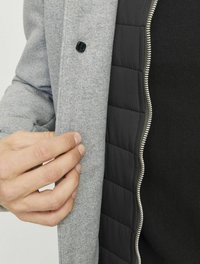 Grau äußere Jacke mit schwarzem, gestepptem Innenfutter, ausgestattet mit einem schwarzen Knopf und silbernem Reißverschluss. Nahaufnahme einer Hand, die den Rand greift.
