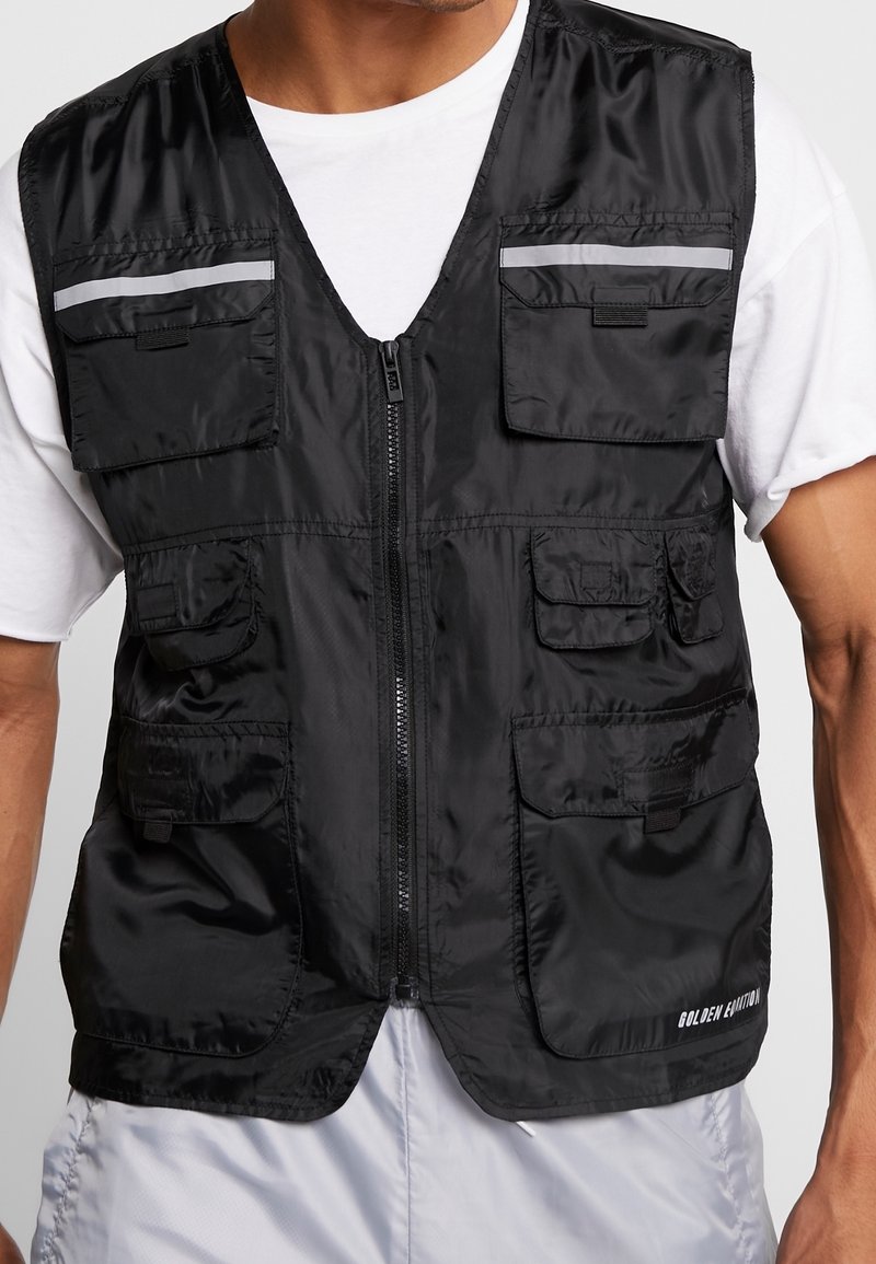 Hombre con chaleco utilitario negro con cremallera y múltiples bolsillos con solapa sobre una camiseta blanca y pantalones gris claro, con el texto "GOLDEN EDITION" en el chaleco.