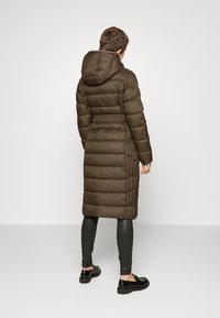Piumino lungo marrone con cappuccio, vita aderente e design trapuntato orizzontale. Abbinato a leggings neri e scarpe.