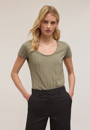 T-shirt vert olive à manches courtes avec un col rond, présentant des motifs de lignes métalliques verticales. Associé à un pantalon noir ajusté.