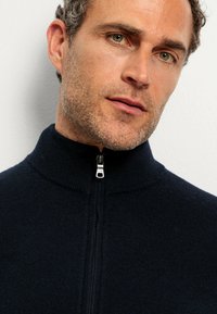 Navy zip-up sweater with a high collar, featuring a metal zipper and ribbed texture. Close-up view highlighting fabric details and design.