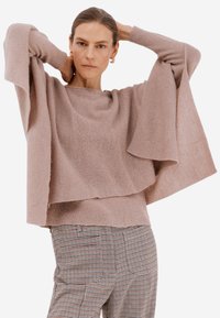 Světlé růžové pletené svetr s širokými rukávy a vrstveným designem, spárovaný s kalhotami s kostkovaným vzorem a kapsami.