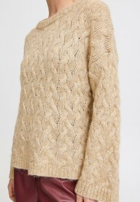 Vaalean beige cable-knit neulepusero, jossa on löysä mitta, leveä pääntie ja pitkät hihat. Pehmeä rakenne, jossa on ribbireunus ja yksityiskohtainen kuvio.