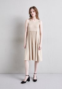Vestido midi de canalé beige con falda acampanada, diseño sin mangas y textura sutil. Combinado con zapatos de tacón cuadrado de ante negro.