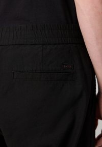 Gros plan d'une personne portant un pantalon noir à taille élastique avec une poche passepoilée à l'arrière et un petit écusson du logo BOSS au-dessus de la poche.