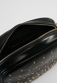 Černá kožená taška s pozlacenými cvočky a zipovým uzavřením. Uvnitř je zelená kapsa s logem značky. Hladký povrch, obdélníkový tvar.