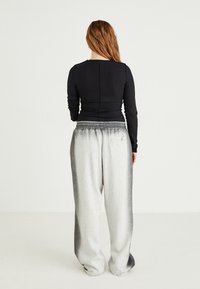 Eine Frau mit langen Haaren steht, trägt ein eng anliegendes schwarzes Langarmoberteil und lockere graue Jogginghosen und ist mit dem Rücken zu einem schlichten weißen Hintergrund abgewandt.