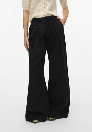 Pantalons larges noirs avec des plis, fabriqués dans un tissu léger. Présentent une taille haute et une coupe décontractée. Accompagnés de sandales vertes.