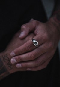Anello d'argento con un design a raggi di sole che presenta un centro nero, indossato su una mano con tatuaggi visibili e dita intrecciate.