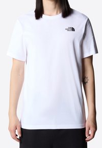 Person wearing a white short-sleeve T-shirt with "The North Face" logo on the chest and a black tattoo on the right forearm.