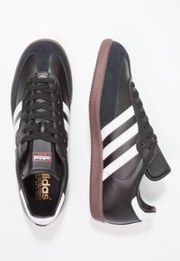 Sneakers Adidas Samba neri con strisce bianche, suole marroni e lacci, mostrati in vista dall'alto e di lato su uno sfondo bianco.