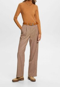 Femme portant un haut à manches longues beige, un pantalon large taille haute à carreaux marron, une ceinture beige, des bottes beiges épaisses et un collier chaîne en or.