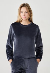 Top lungo in velluto blu navy a maniche lunghe con texture a coste, scollatura rotonda e polsini arricciati. Abbinato a pantaloni coordinati, progettato per il comfort.