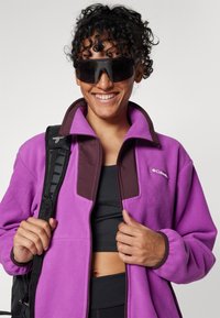 Purple fleece jacket with a brown collar, featuring a zipper and logo. Model wearing dark sunglasses and holding a black backpack strap.