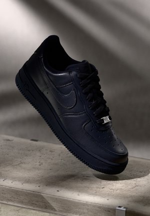 Schwarzer Nike Air Force 1 Sneaker mit schwarzen Schnürsenkeln und metallischem Schnürsenkelanhänger, schräg auf einer Betonoberfläche mit weichem Schattenwurf positioniert.