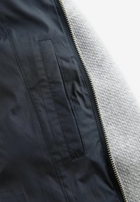 Vue de face d'une veste avec un intérieur en nylon noir, un extérieur en polaire texturée gris, avec fermeture éclair et poche latérale.