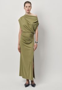 Olijfgroene maxi-jurk met een asymmetrische halslijn, gedrapeerde lijfje, zijsplit en een gladde textuur. Geaccentueerd met zwarte puntige ballerina's.